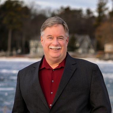 A man in a suit and red shirt is smiling in front of a body of water.