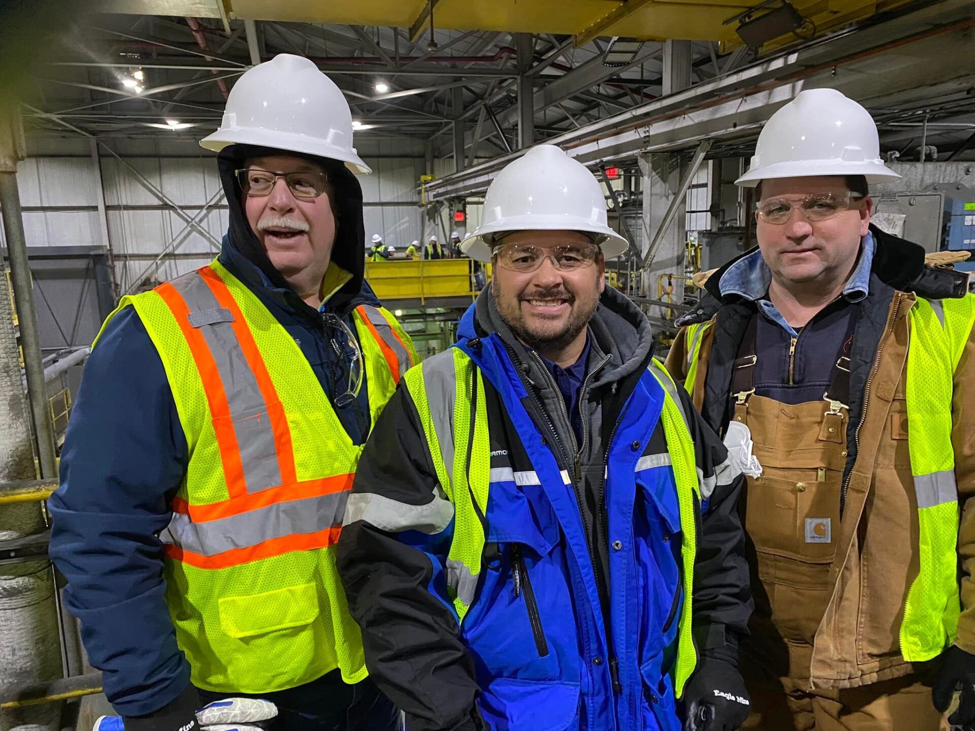 Three men wearing hard hats and safety vests are posing for a picture in a factory.