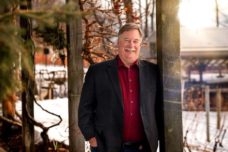 A man in a suit and red shirt is leaning against a tree in the woods.