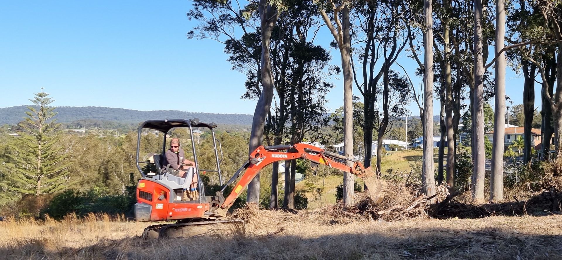 An orange excavator clearing debris near trees on a sunny day. A person is operating it.