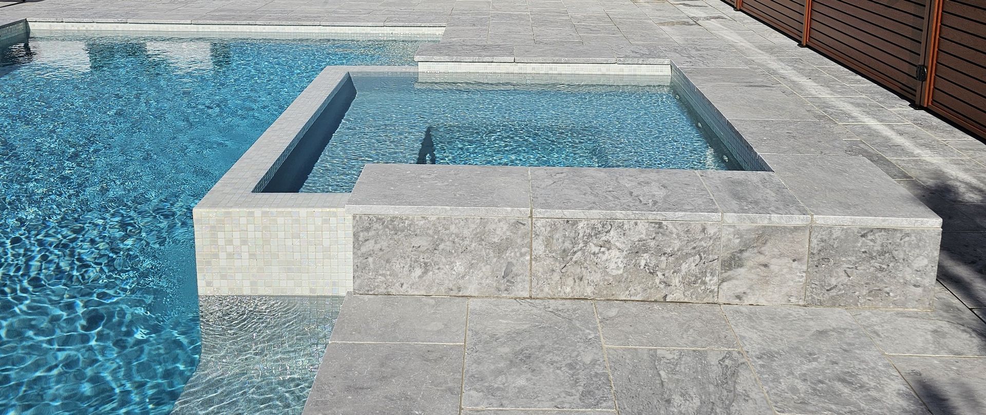 A modern swimming pool with built-in spa. Light gray stone borders both. Wooden fence visible on right.