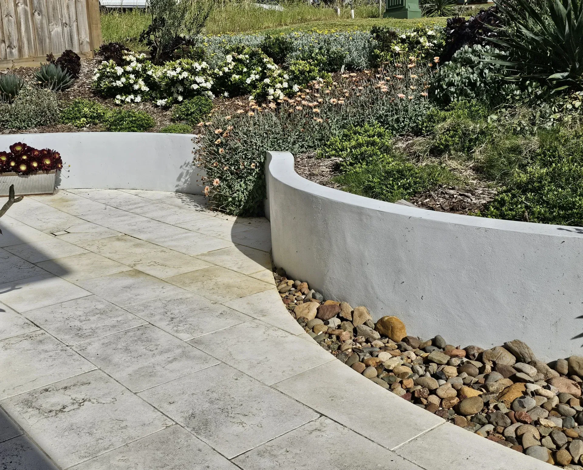 Curved stone path leads past a concrete wall with a garden bed full of plants and rocks.