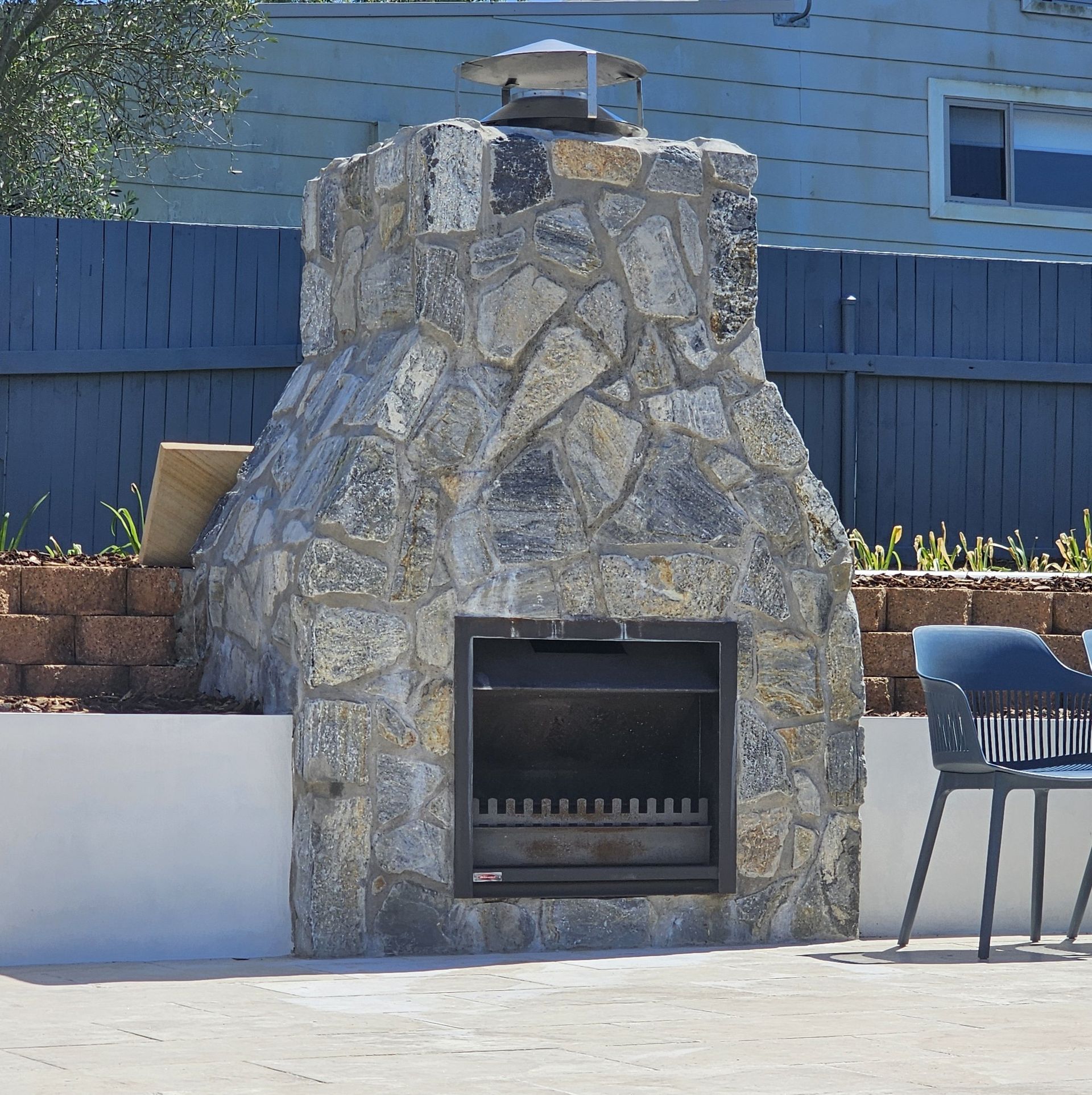 Stone outdoor fireplace with a metal firebox and chimney, in a backyard setting.
