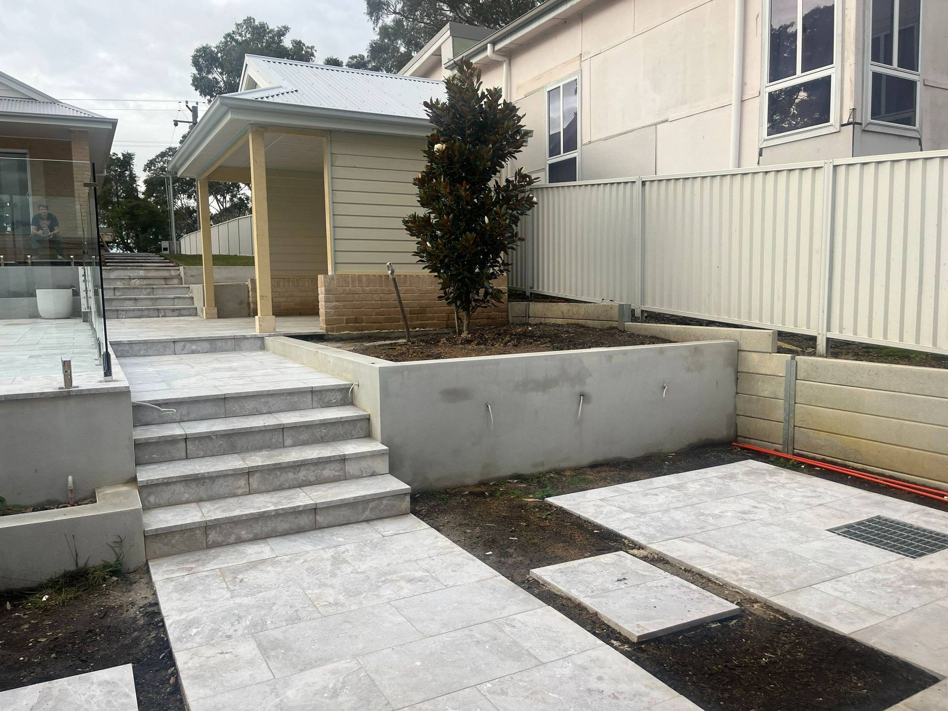 Exterior view of a building with stone steps leading up to a small structure, with a young tree in a raised bed.