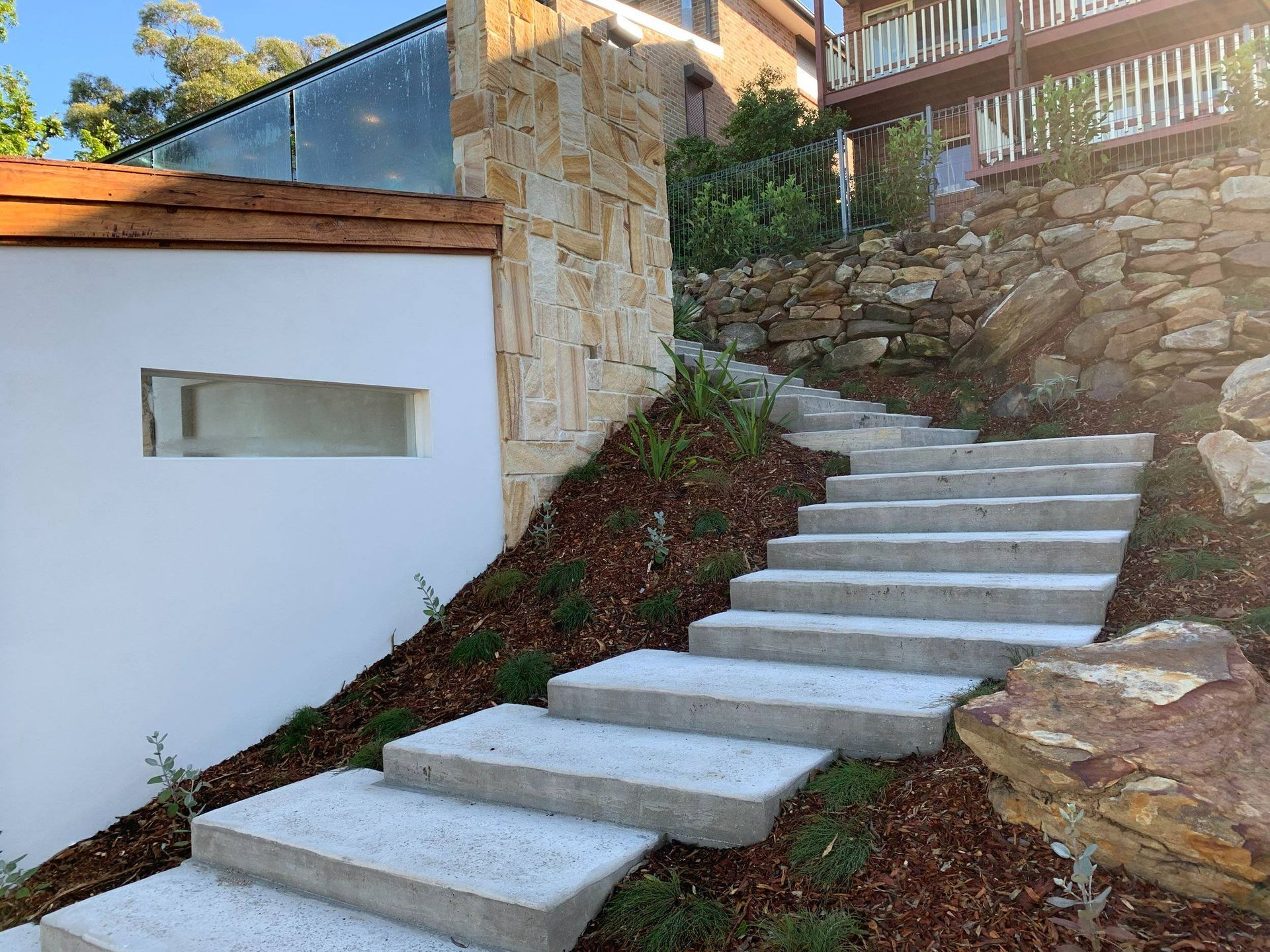 Concrete steps ascend a hillside, flanked by landscaping and a stone wall.