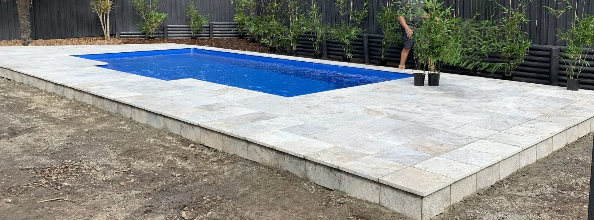 A rectangular swimming pool covered with blue material, surrounded by concrete and dirt. Green plants in background.