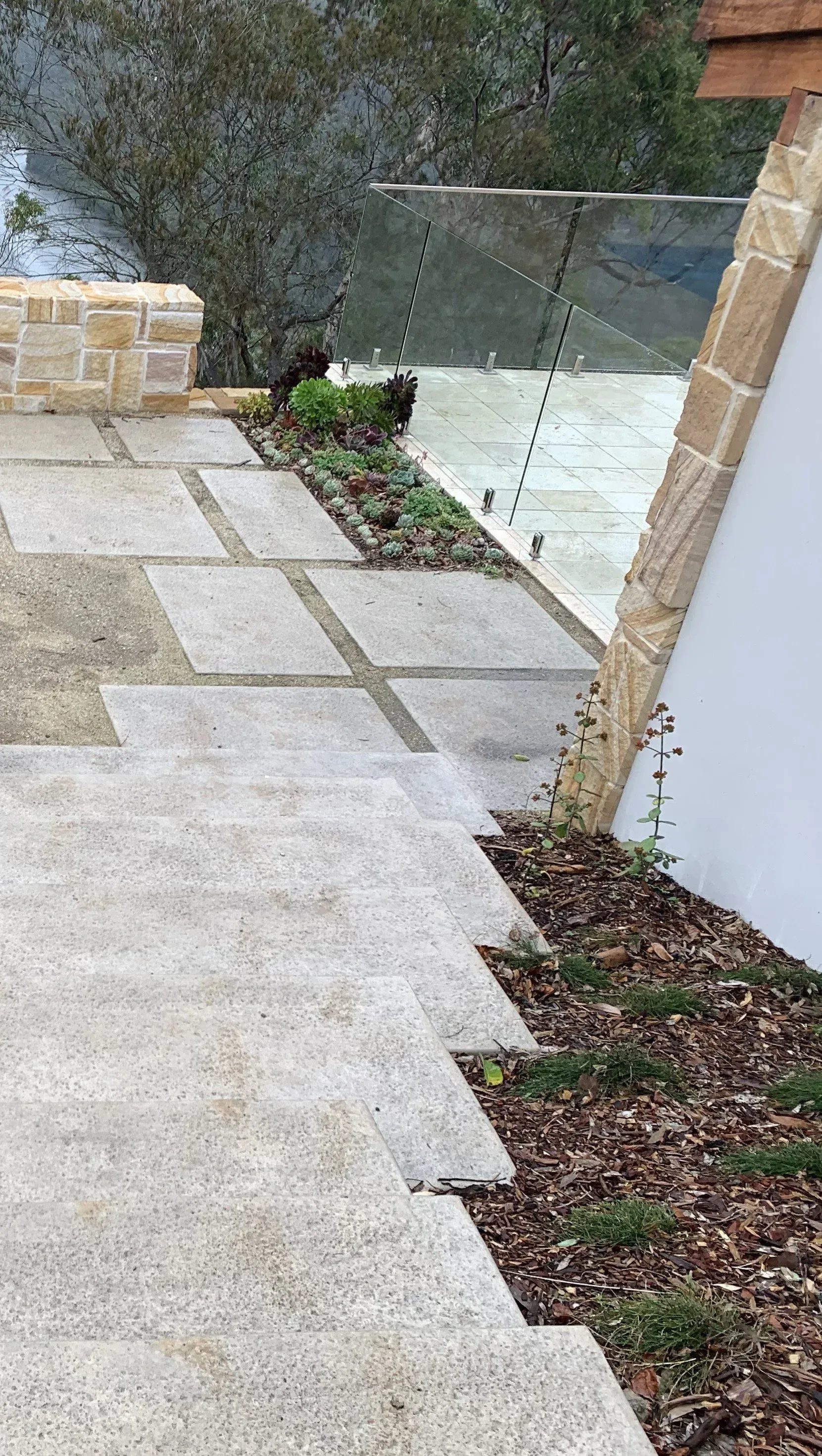 Stone steps and pathway with glass railing, bordered by small plants and wood chips.