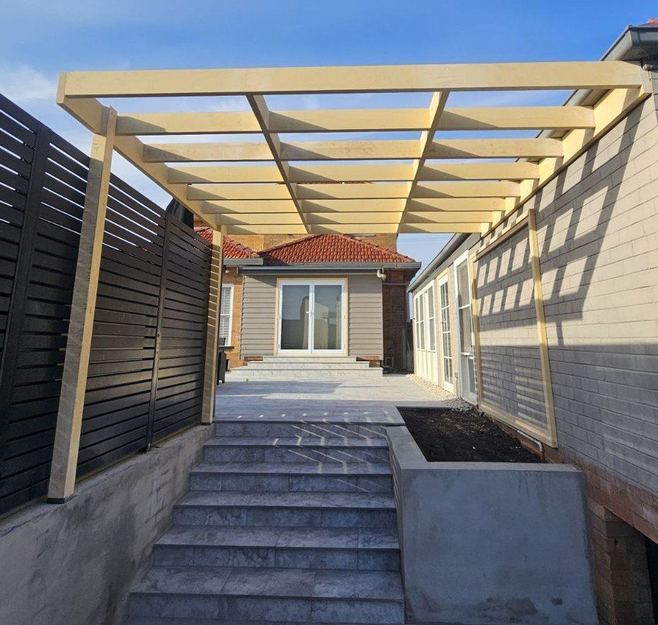 Wooden pergola over concrete steps leading to a house, flanked by a dark fence and building wall.