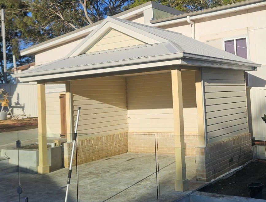 A covered outdoor structure with a light gray metal roof, wooden beams, and beige siding; located near a concrete patio.