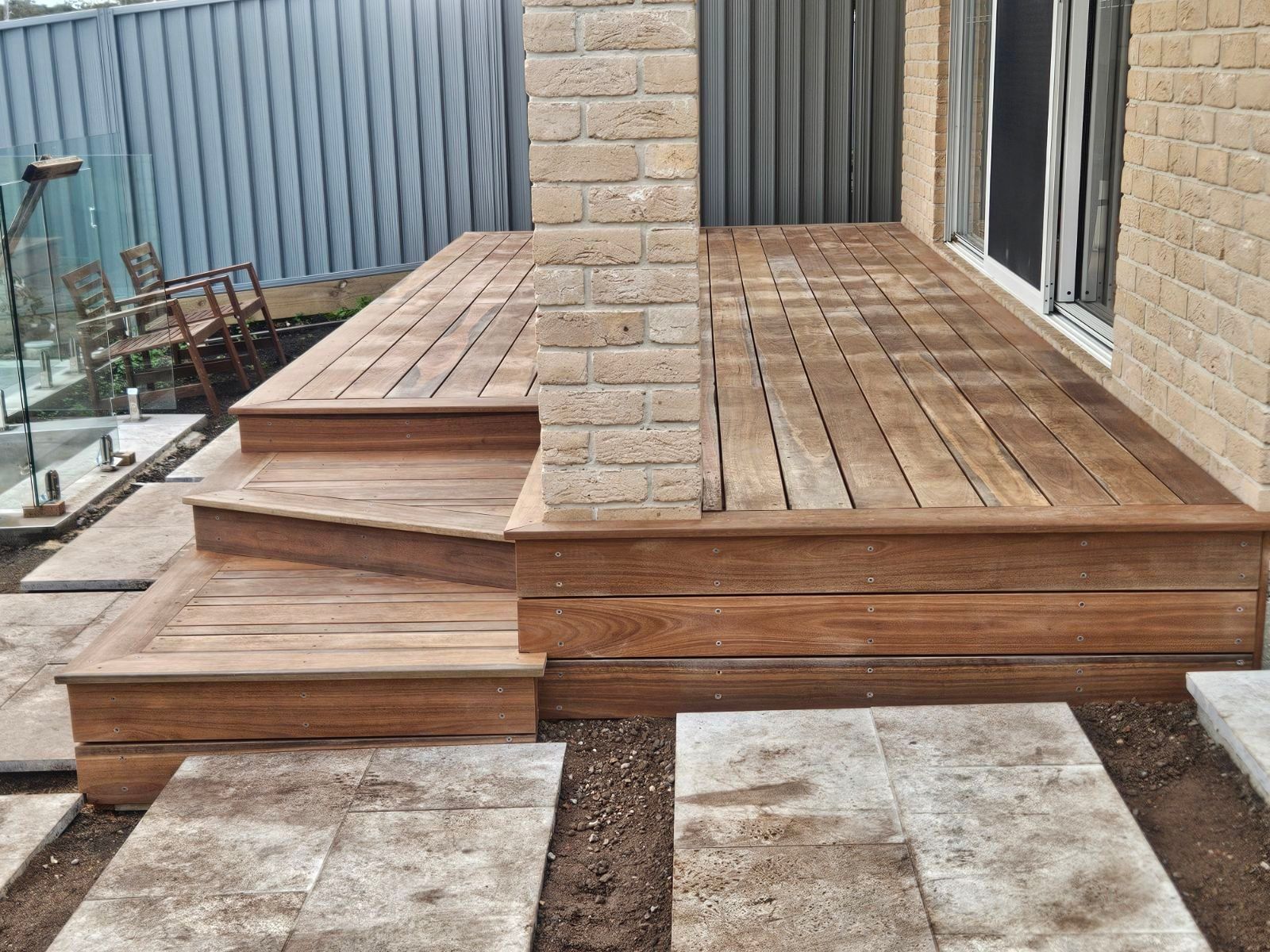 Wooden deck with steps, built against a brick wall, adjacent to a patio with stepping stones.