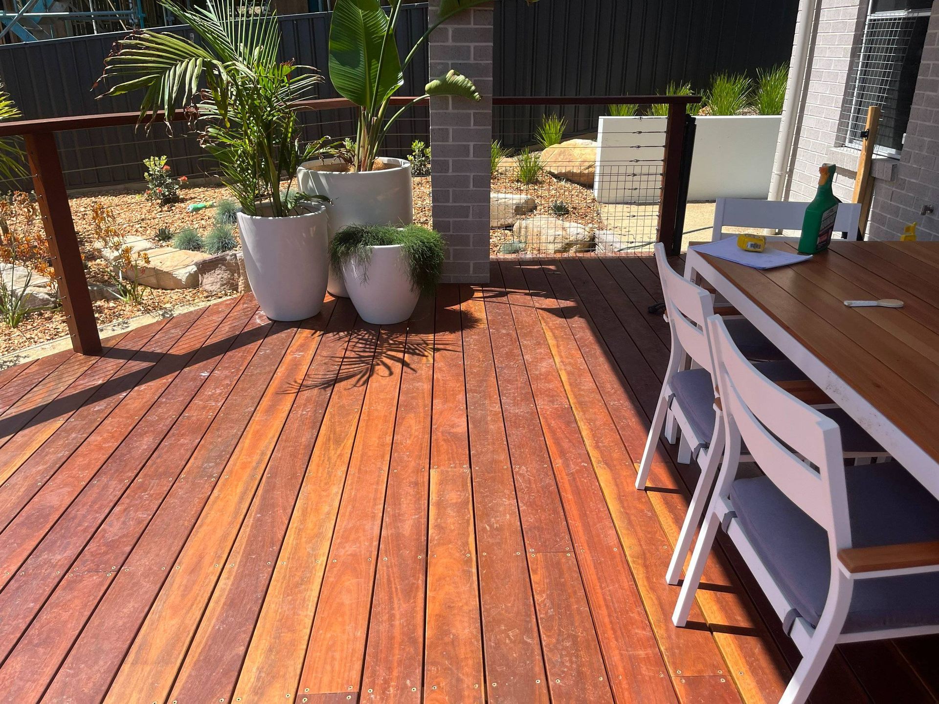 Wooden deck with potted plants, table, chairs, and a bottle; sunny outdoor setting.