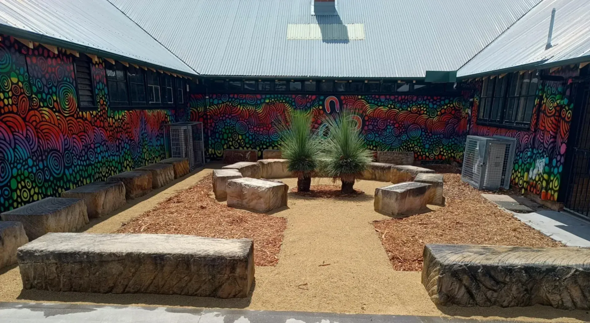 Courtyard with hay bale seating, gravel ground, colorful mural, and two potted plants.