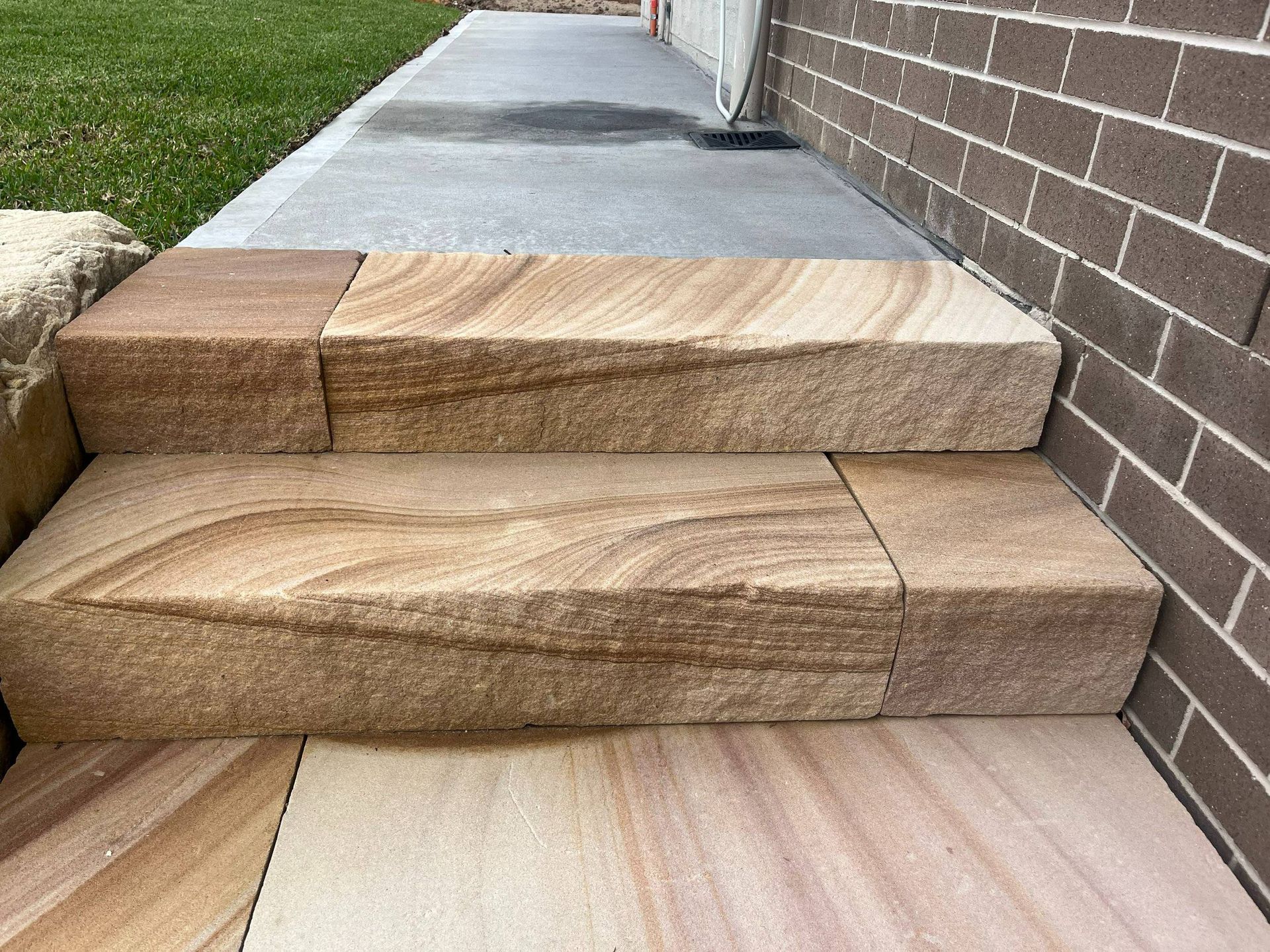 Sandstone steps leading to a concrete surface, next to a brick wall and lawn.