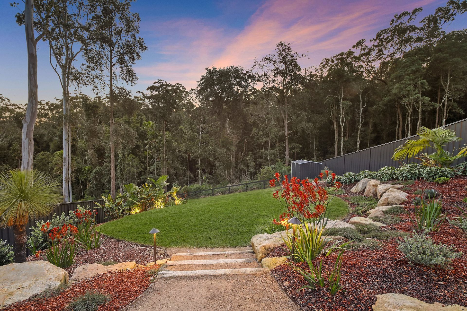 Pathway leading to a grassy hill, surrounded by landscaped gardens and trees, under a twilight sky.