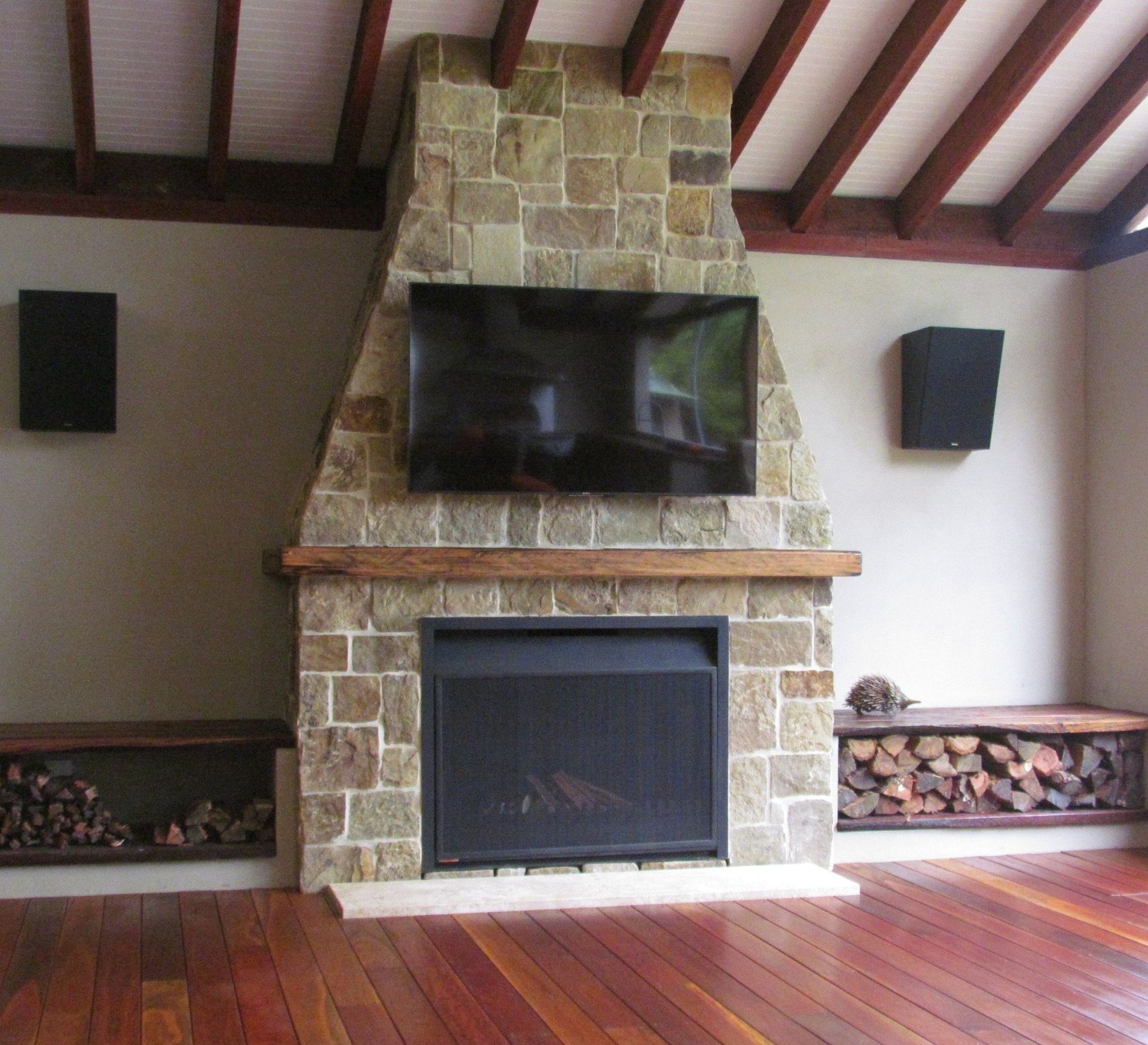 Fireplace with stone facade, TV, and speakers. Wooden benches with firewood on each side.