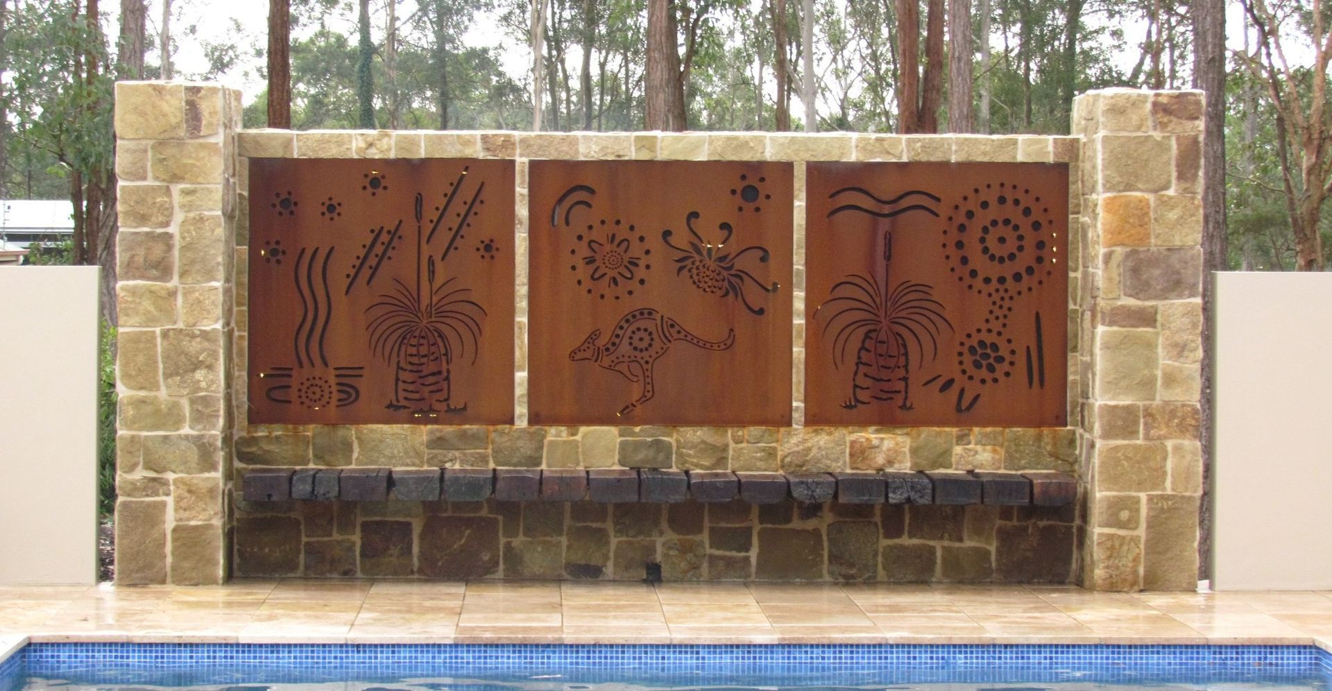 Poolside water feature with three rusty metal panels featuring cut-out botanical designs, framed by stone columns.