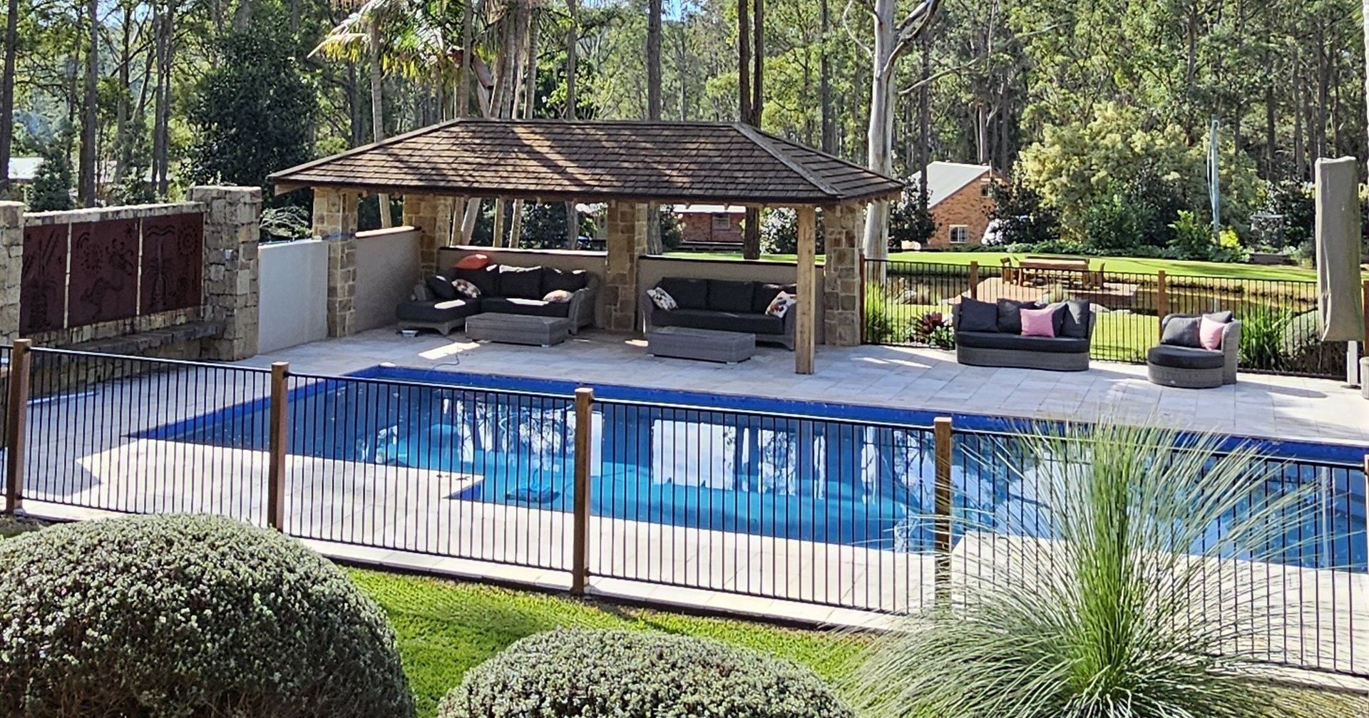 Poolside gazebo with seating, fenced pool, and trees in a sunny outdoor setting.