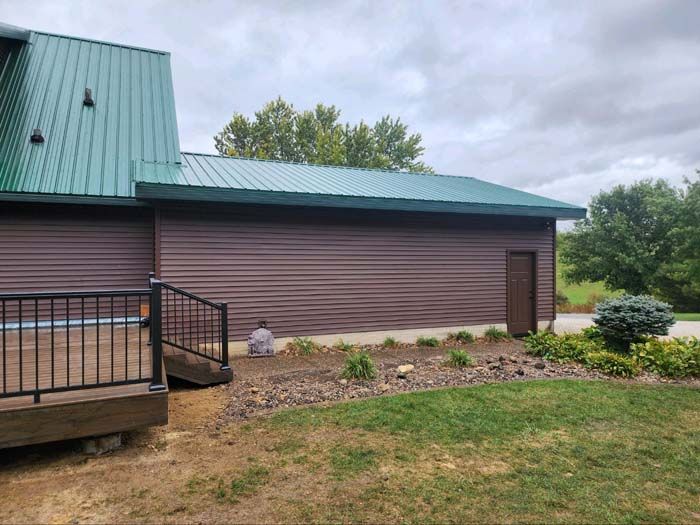 Side of a house with green metal roof, brown siding, porch railing, and a grassy yard with shrubs.