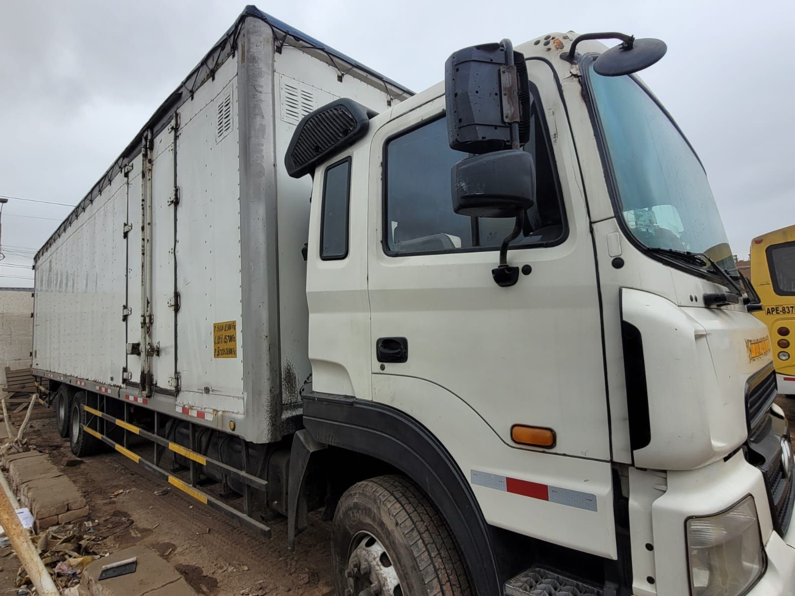 Una fila de furgonetas blancas aparcadas en el terreno de un almacén, con un gran camión de transporte al fondo.