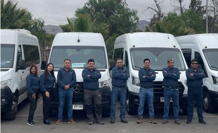 Trabajadores de pie frente a unas furgonetas blancas en un aparcamiento, con los brazos cruzados y uniformes azules.