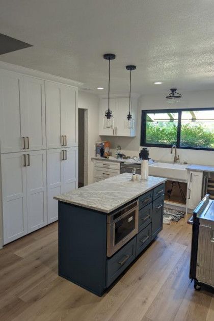 Modern kitchen with white cabinets, dark island, marble counters, pendant lights, and large window.