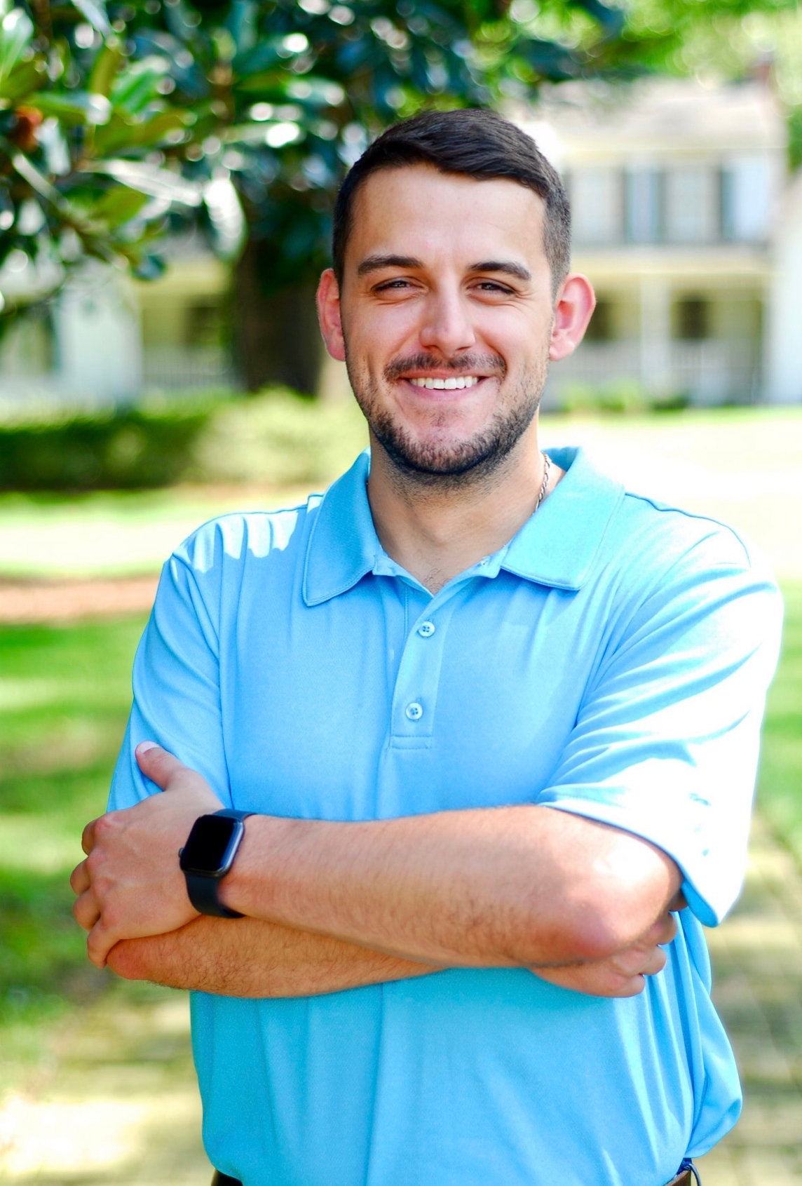Man in a button-down shirt smiles, leaning against a wooden post. Floral arrangement in background.