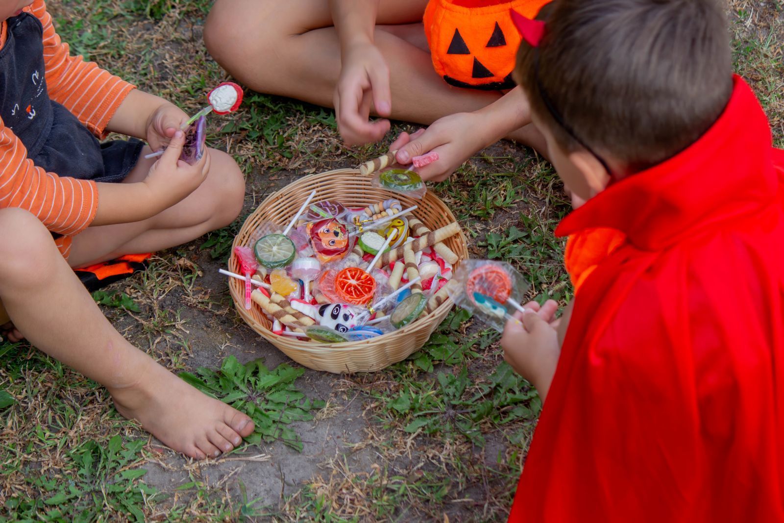 Children gathered around a Halloween treat basket outdoors, with pumpkins and snacks on the grass.