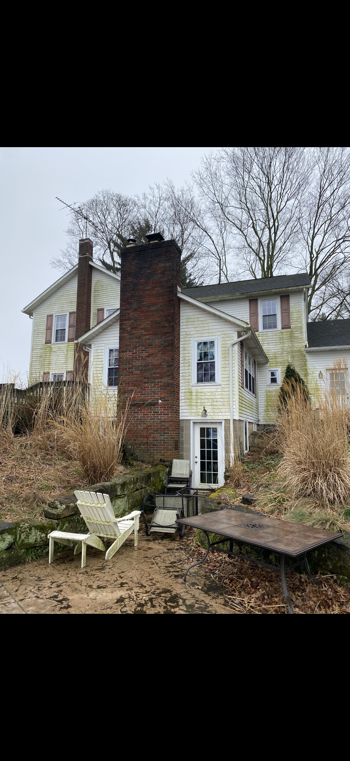 Weathered white house with red brick chimney, overgrown yard, and a white lawn chair in front.