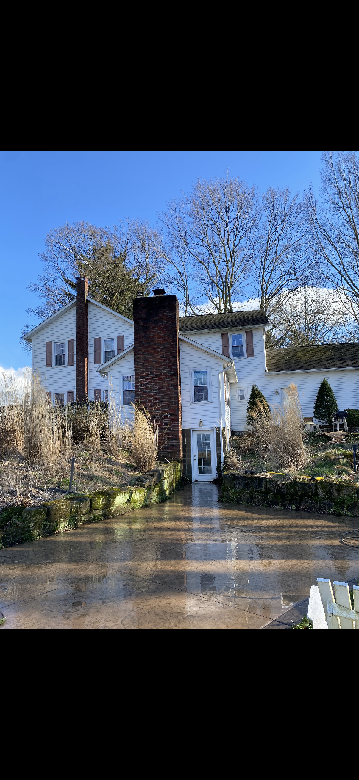White house with brick chimney by a wet driveway, trees overhead, and a clear blue sky.