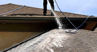 Worker pressure-washing a roof, spraying water across brown shingles with a hose.