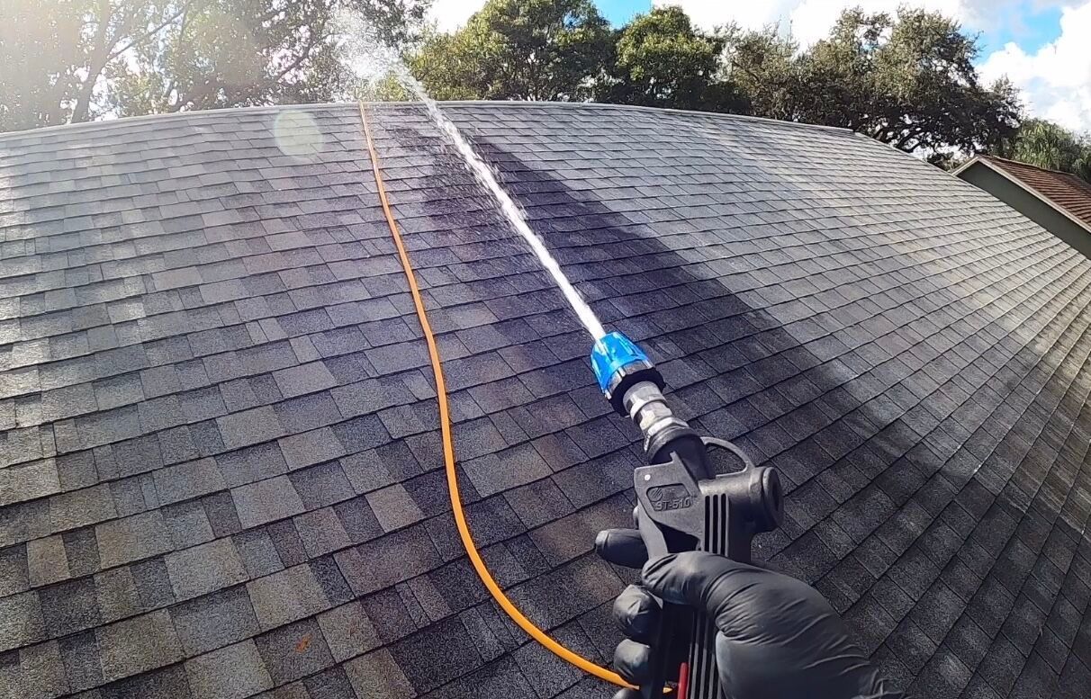 Worker pressure-washing a dark shingle roof with a long spray wand and orange hose