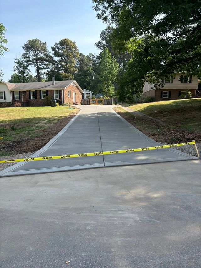 Concrete driveway blocked by yellow caution tape, leading to a brick house with trees and grass on both sides