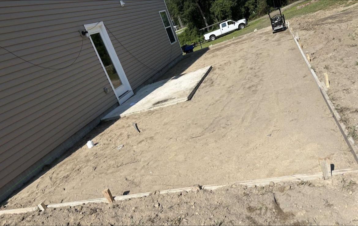 Tilted view of a sandy yard beside a house, with a concrete step and small posts in the ground