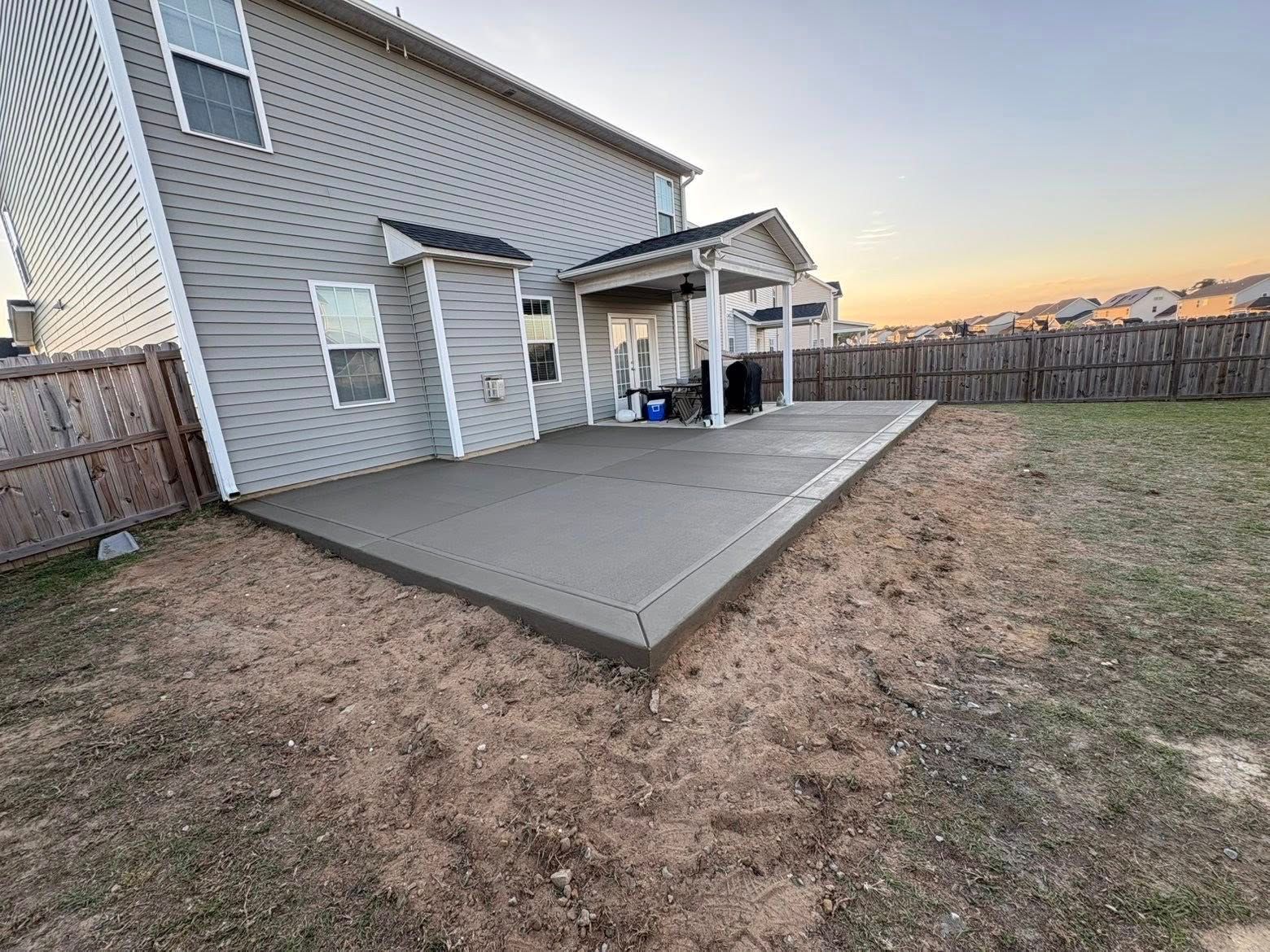 Backyard concrete patio beside a beige house at sunset, with a covered grill area and fenced yard.