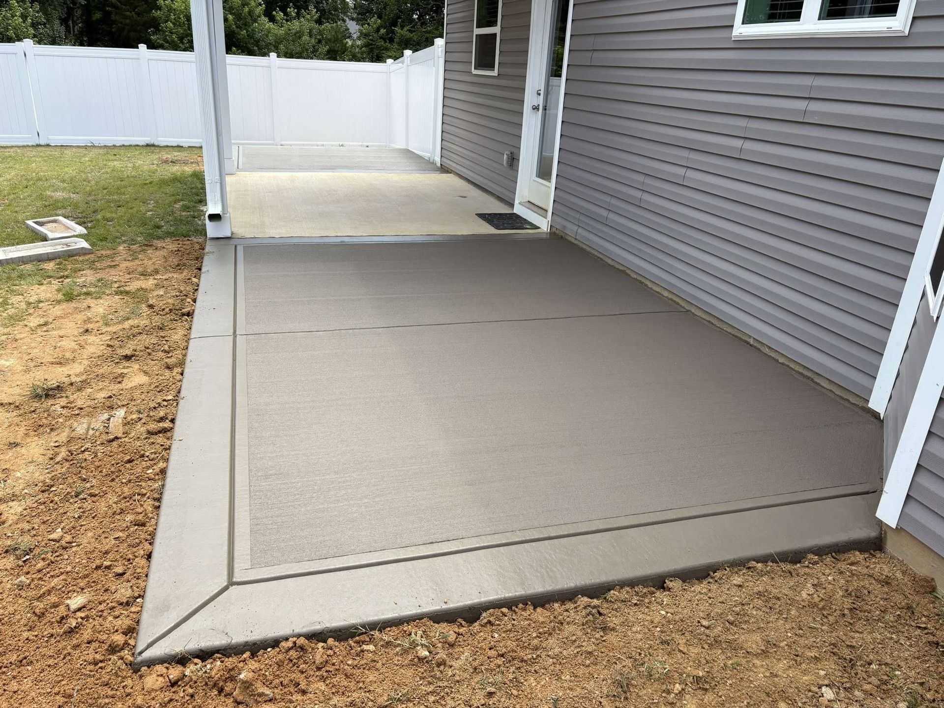 Concrete patio beside gray house, bordered by grass and white fence gate.