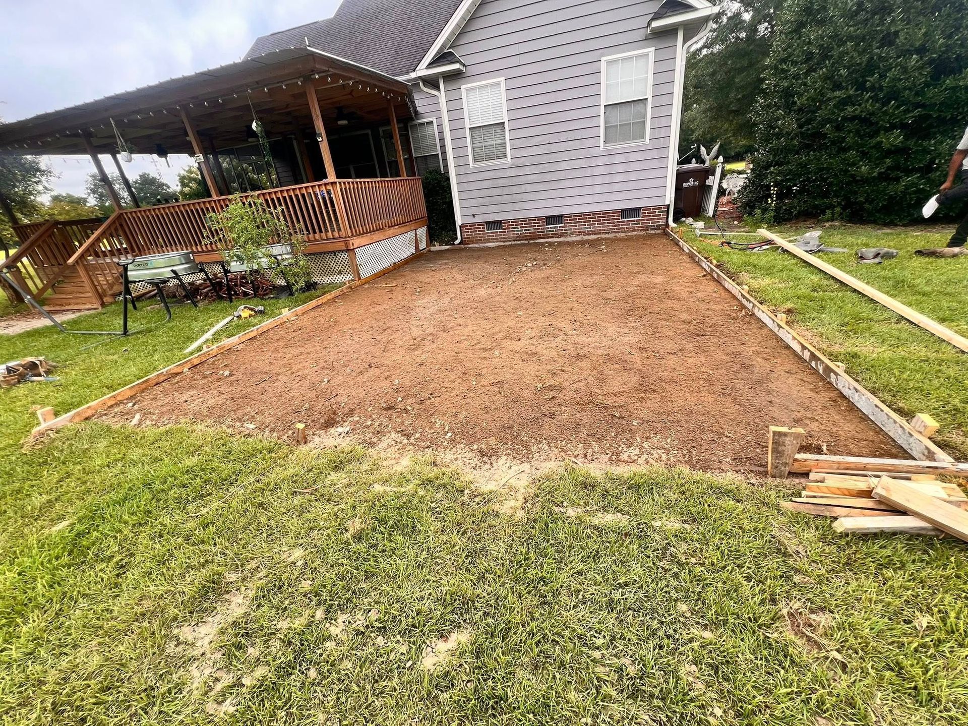 Freshly graded dirt area beside a house and covered patio, with grass and stacked boards nearby