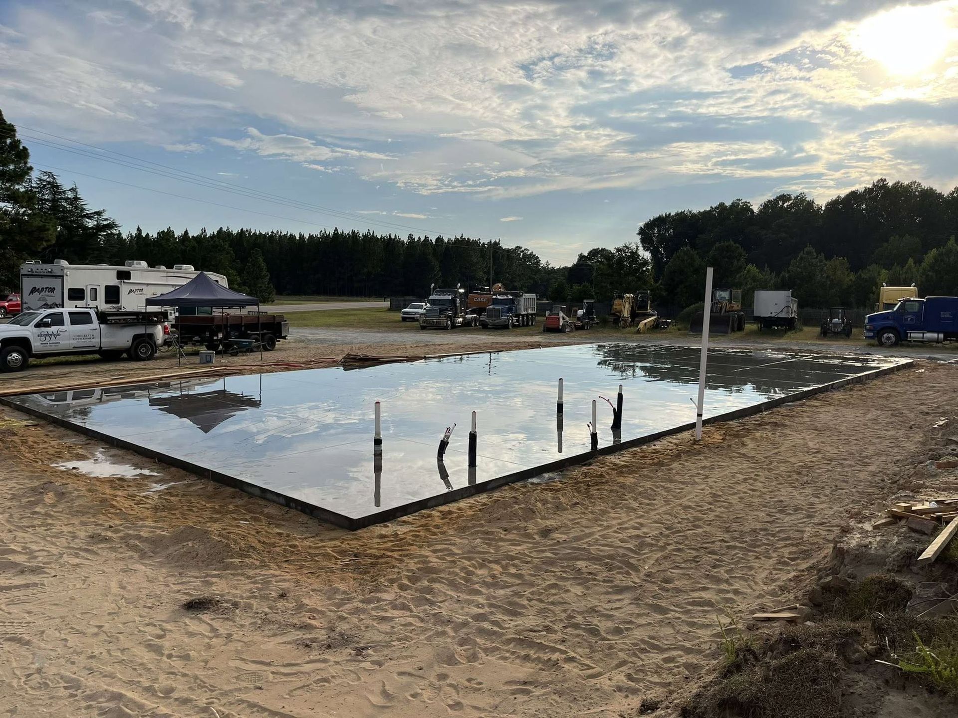 Construction site with a concrete foundation slab, wet surface, and trucks under a cloudy sky