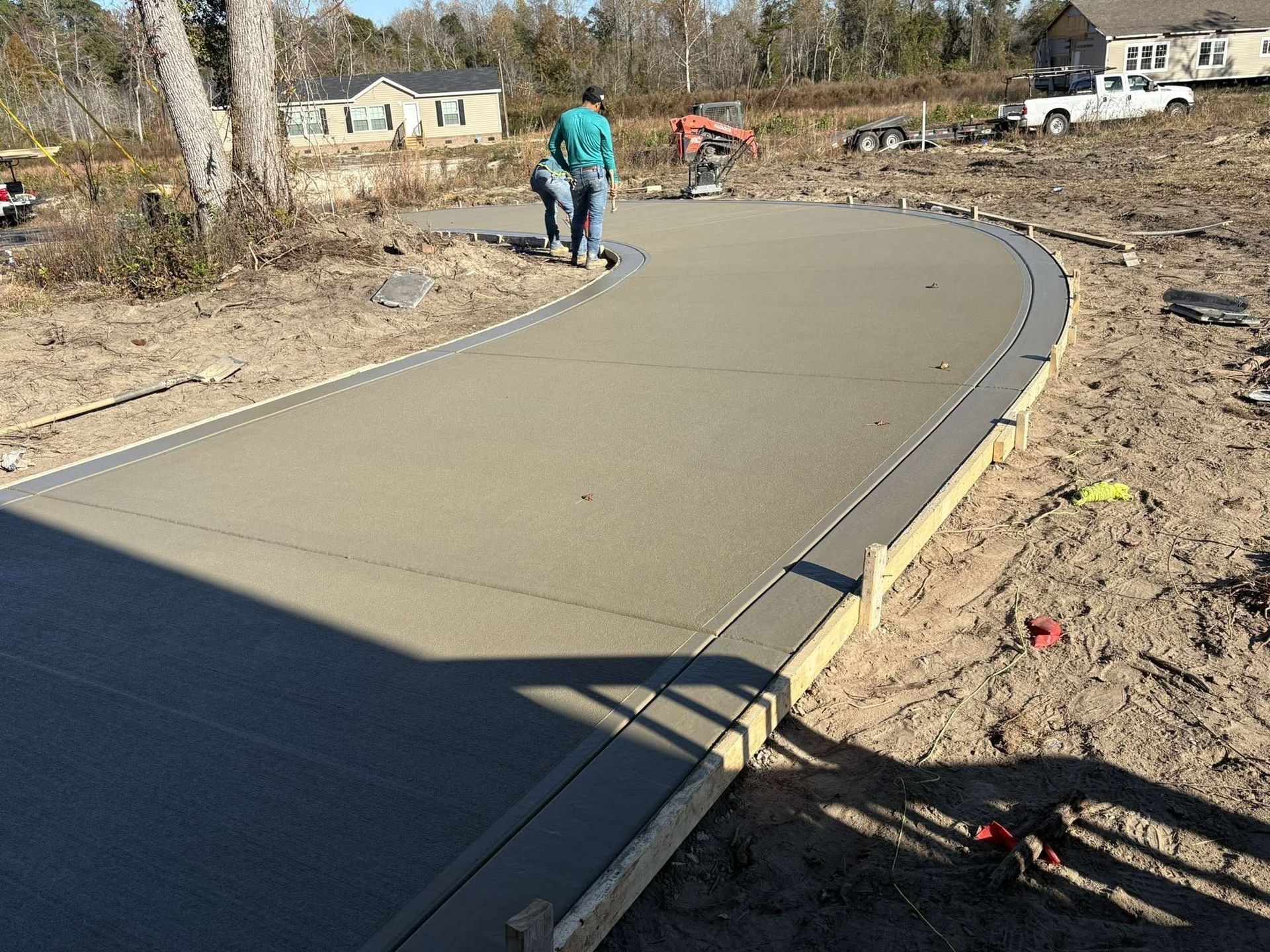 Curving concrete path under construction, with worker, dirt edges, and houses in the background