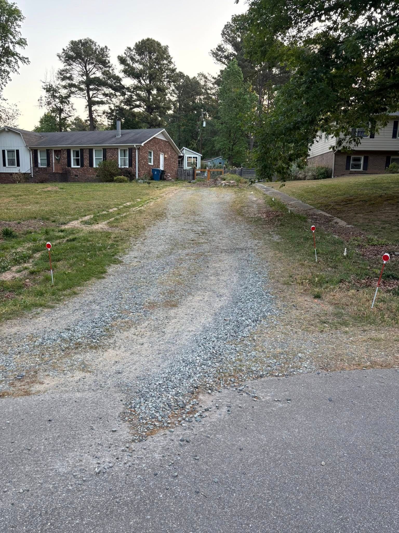 Gravel driveway leading to a house on a grassy lot, with trees and a parked trailer in the background