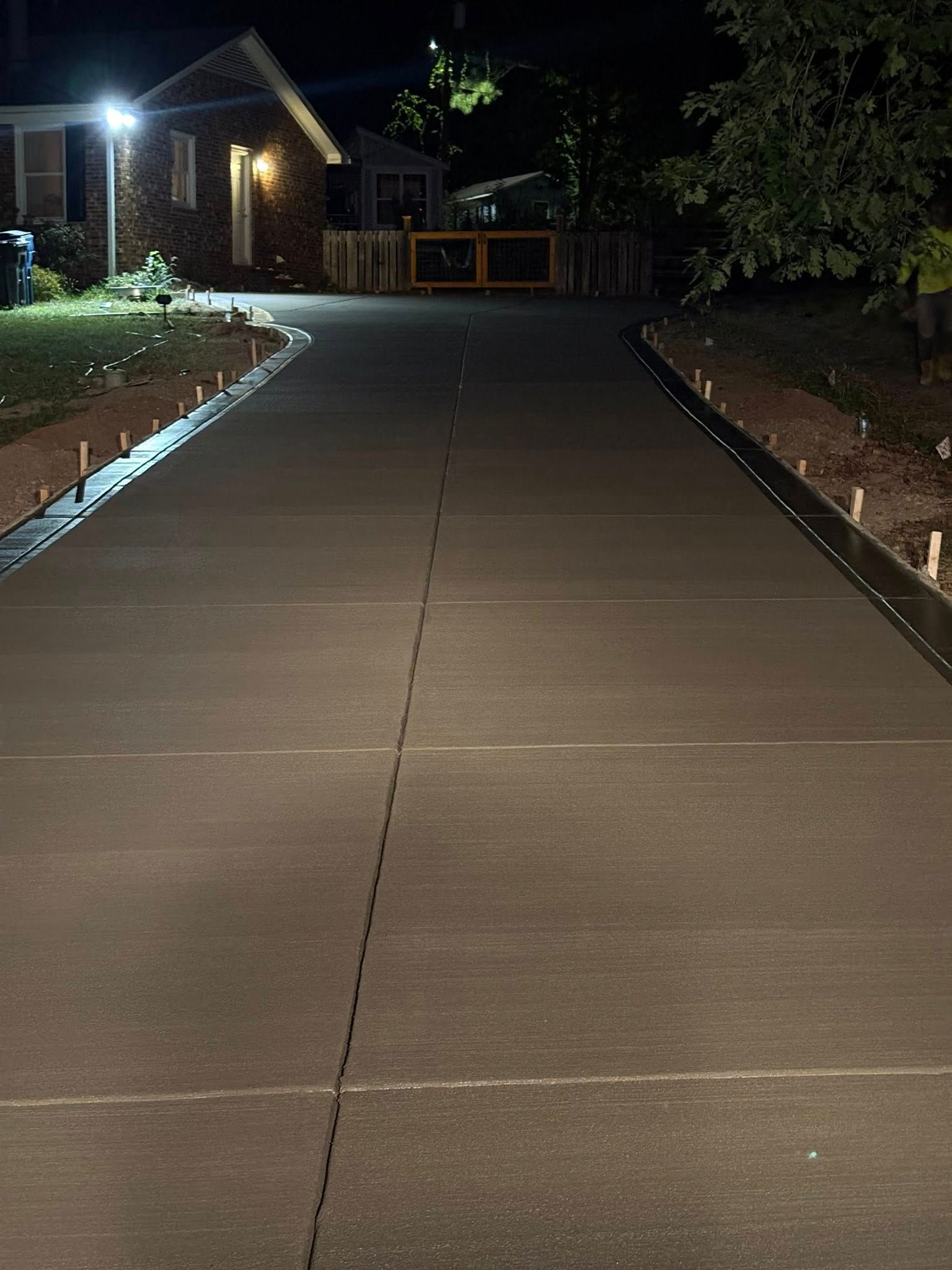 Empty concrete driveway at night beside a brick house, lit by a porch light and surrounded by trees.