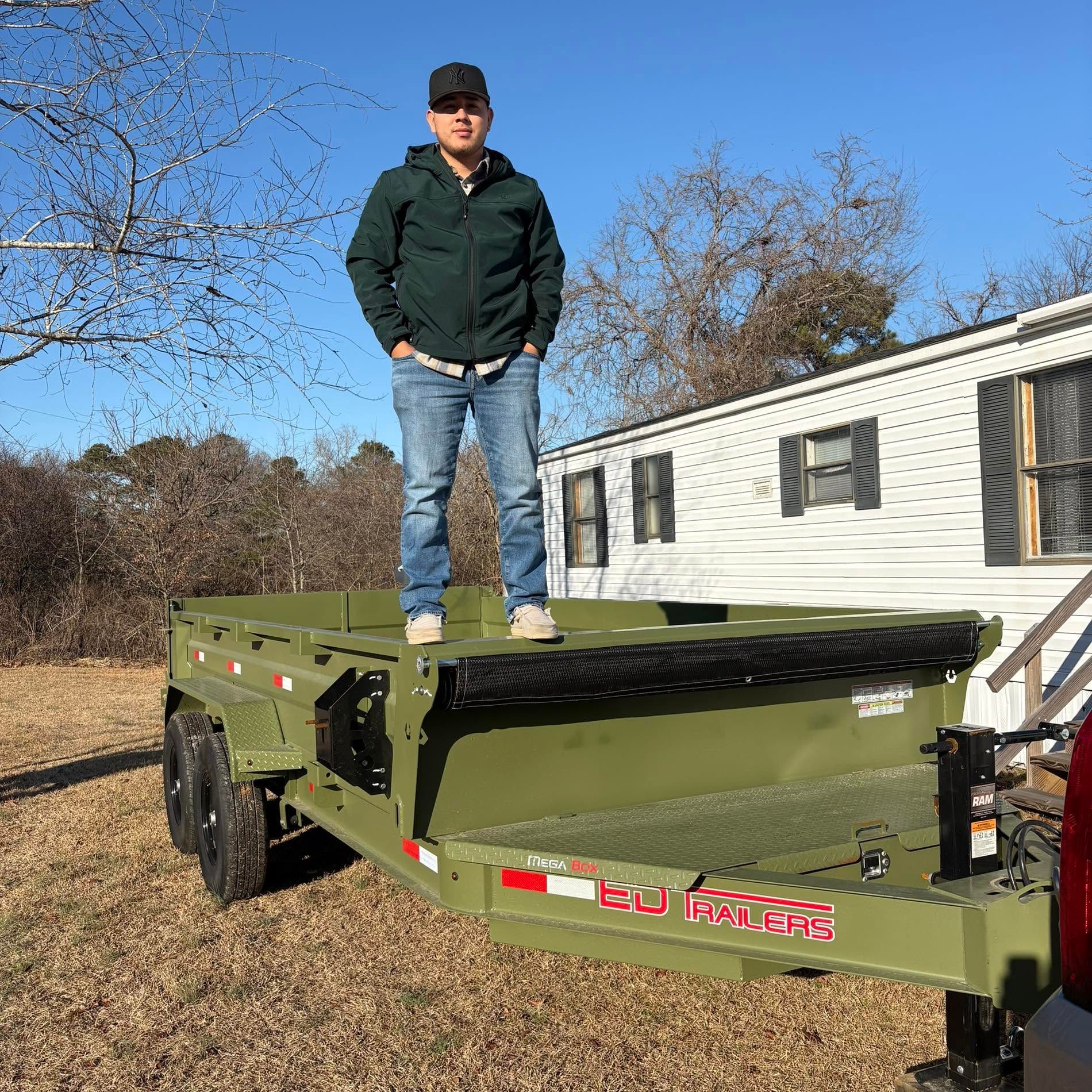 Man standing on a green utility trailer beside a white mobile home on a sunny day