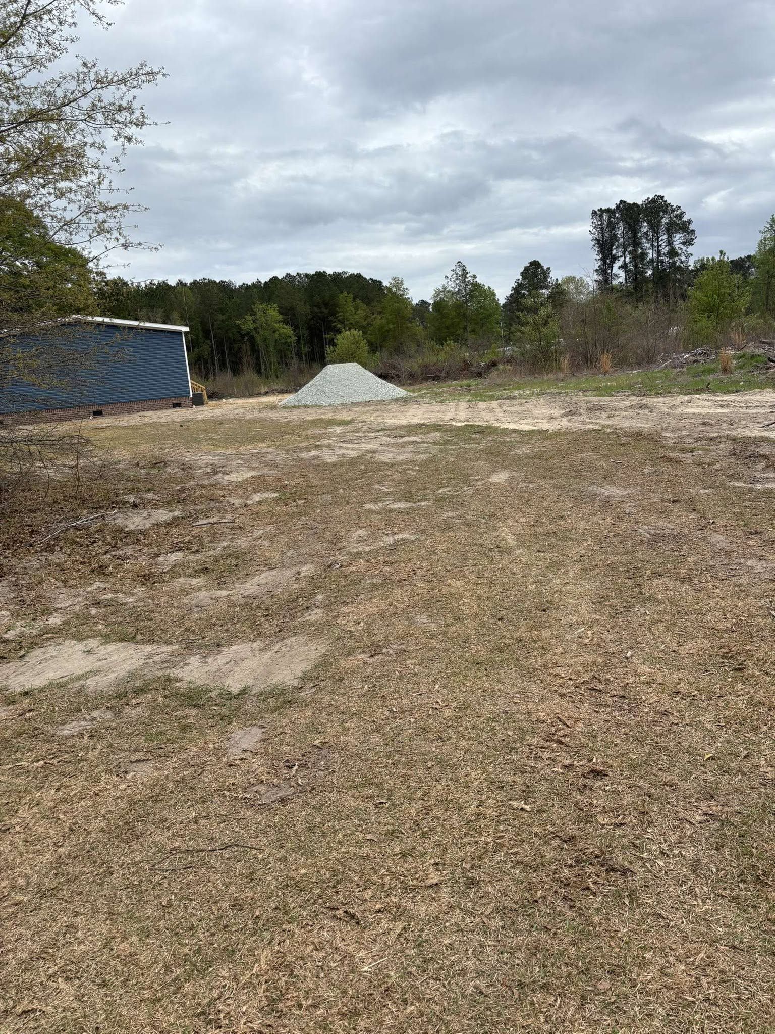 Gravel lot with a pile of sand, a blue structure on the left, and trees under a cloudy sky.
