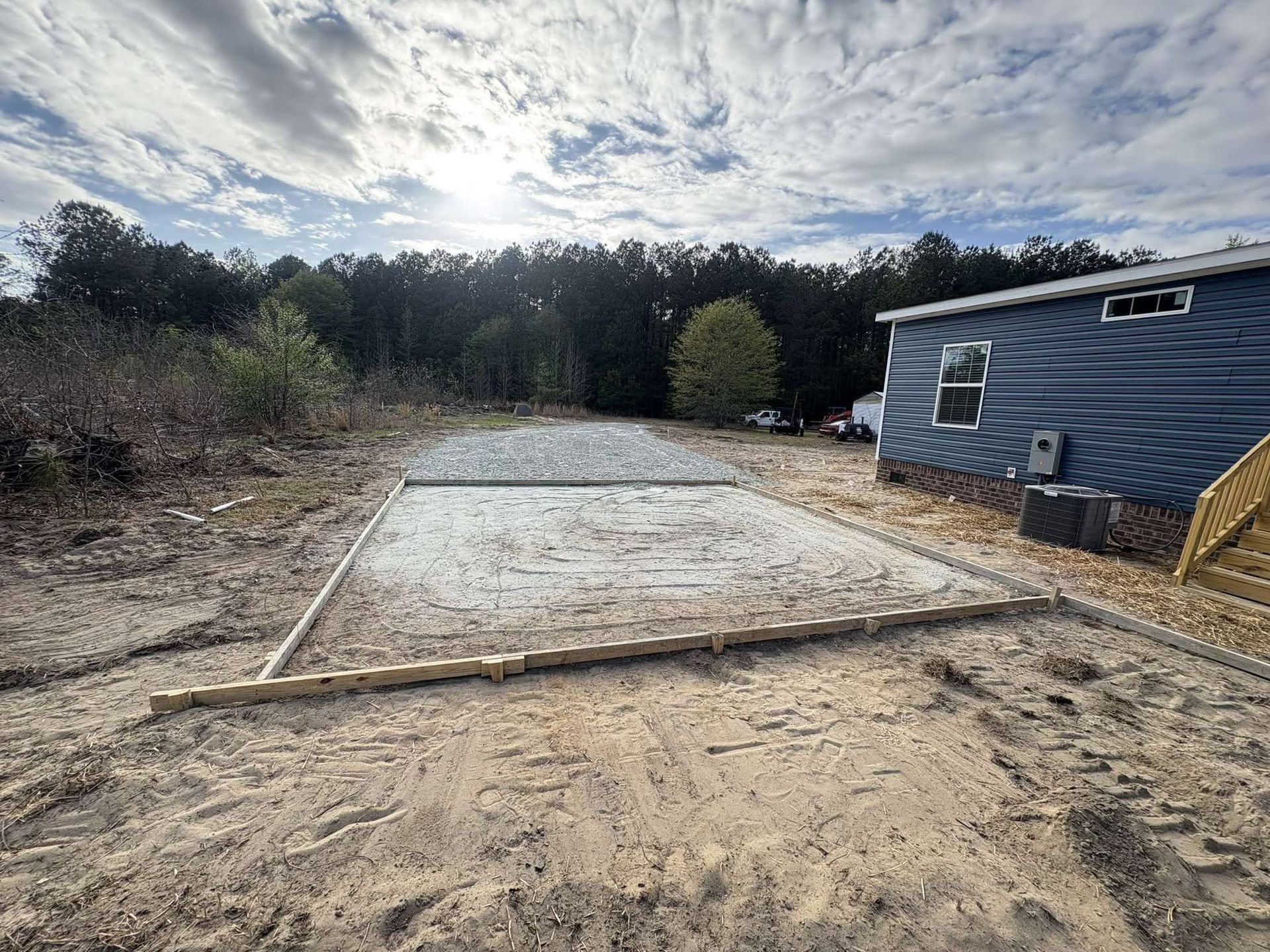 Concrete slab foundation site beside a blue mobile home on a sandy lot with trees in the background