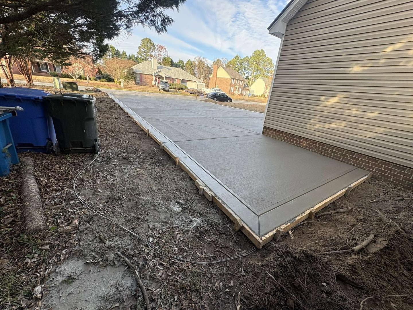 Concrete side ramp beside a house, bordered by dirt and trash bins in a residential yard.