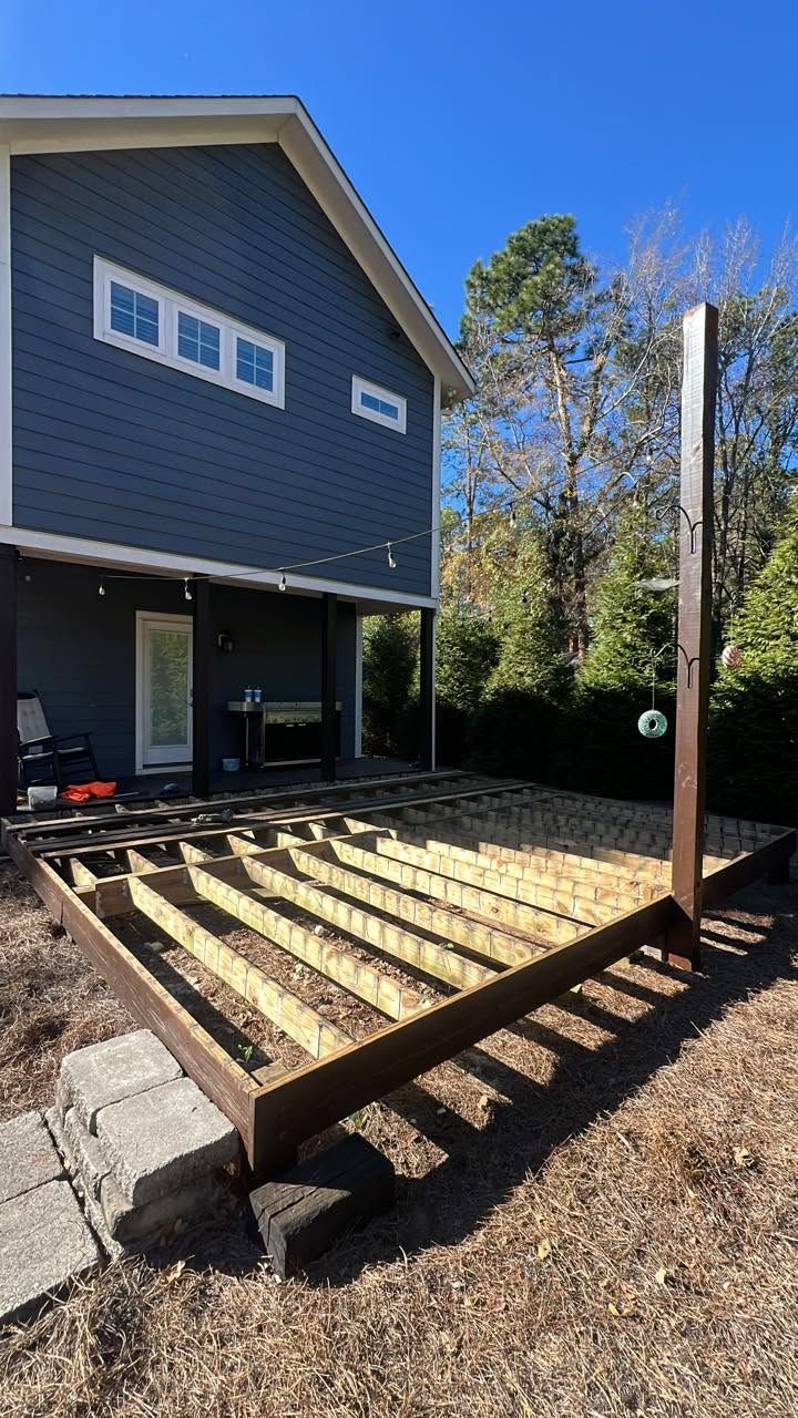 Backyard deck framing beside a dark blue house, with a tall support post and gravel ground