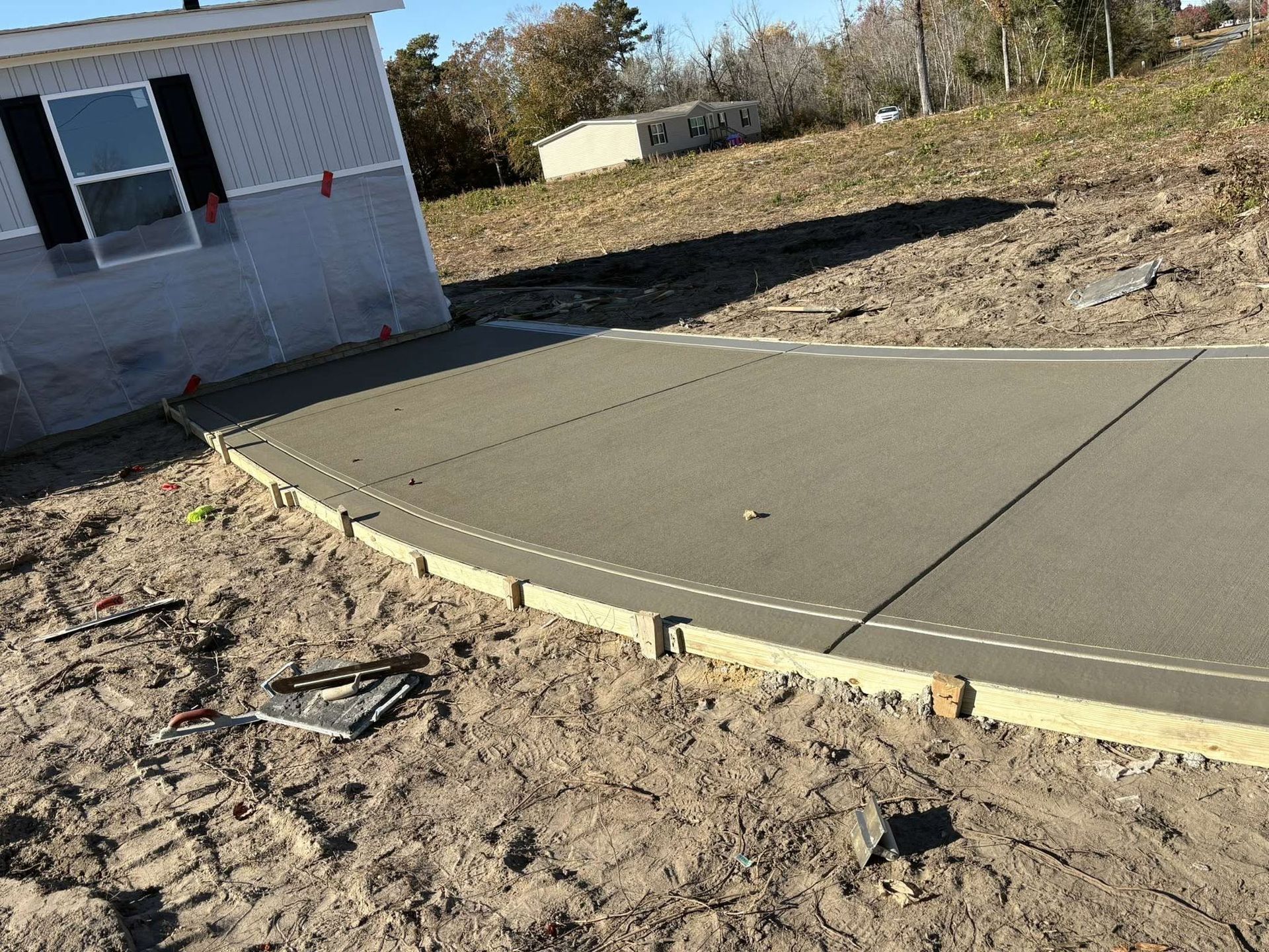 Curved concrete patio beside a house, bordered by dirt and a grassy yard.