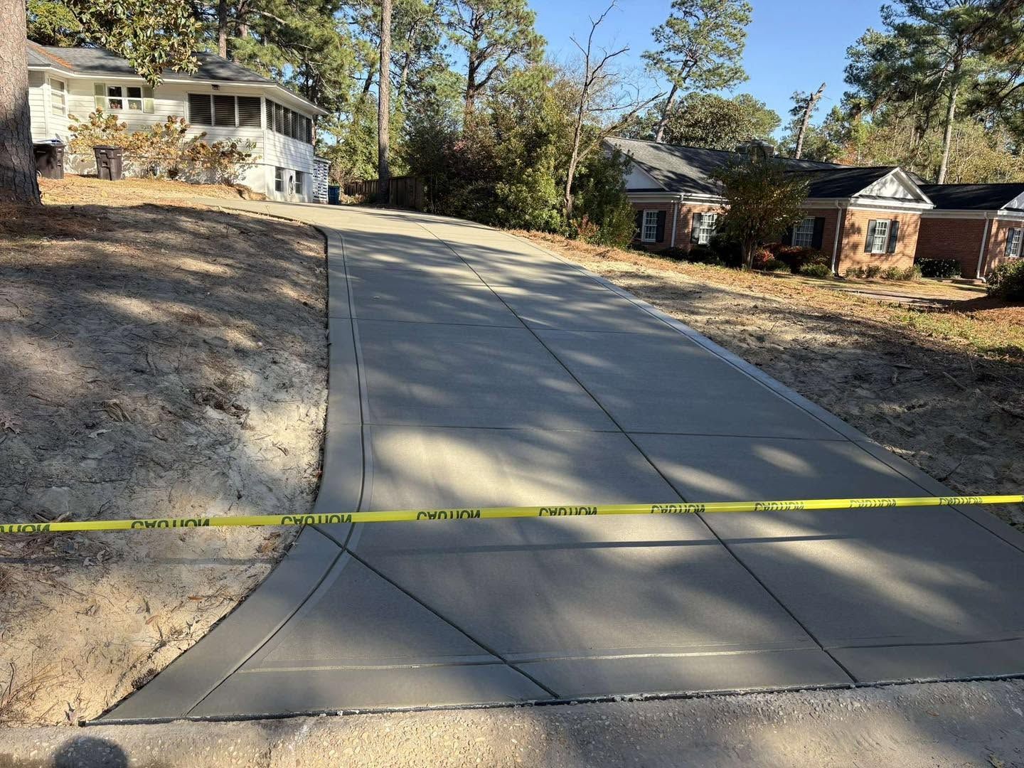 Steep concrete driveway under construction, cordoned off with yellow caution tape beside houses and trees.