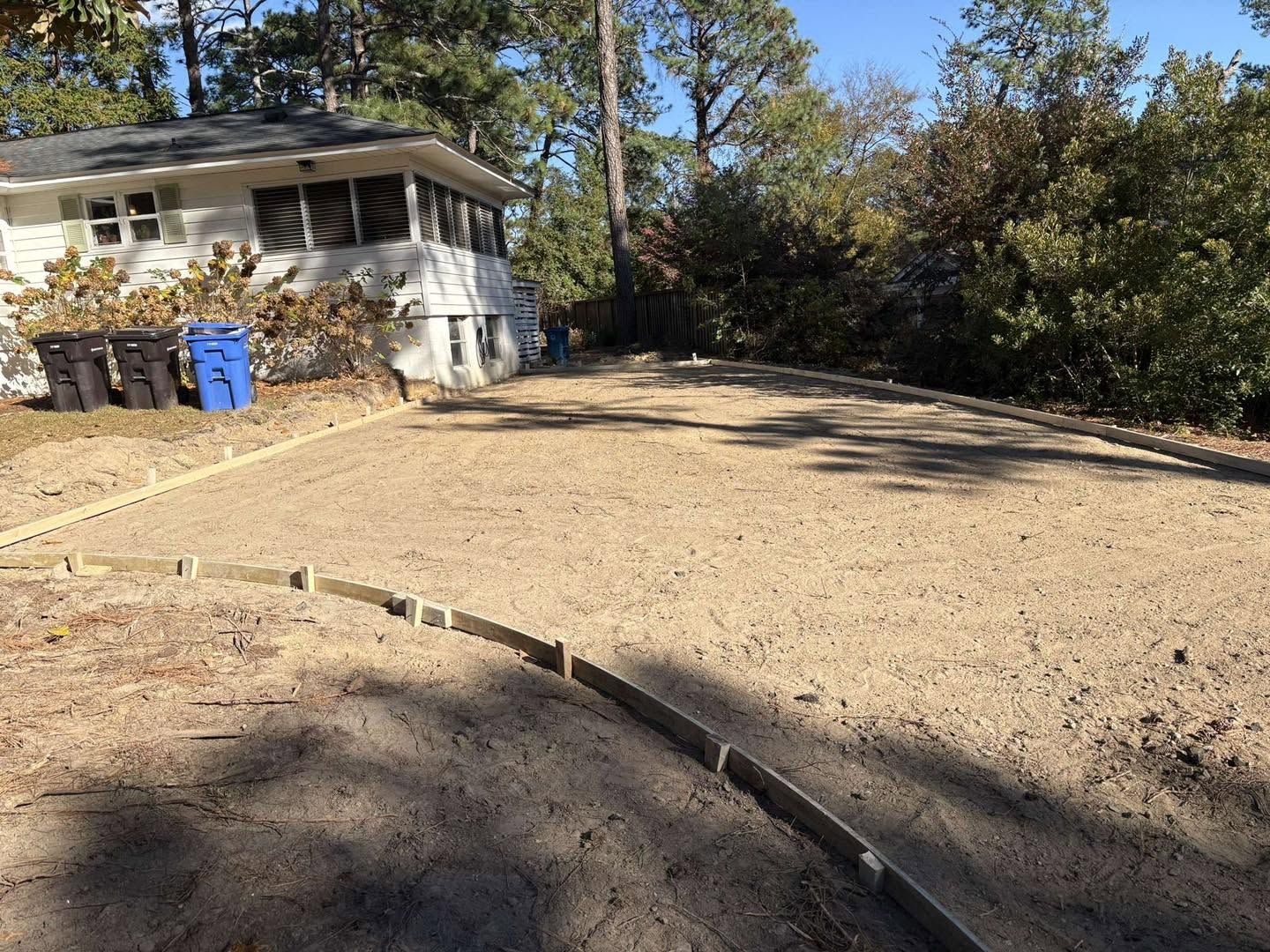 Driveway and front yard with a small house, two blue bins, and trees in bright sunlight