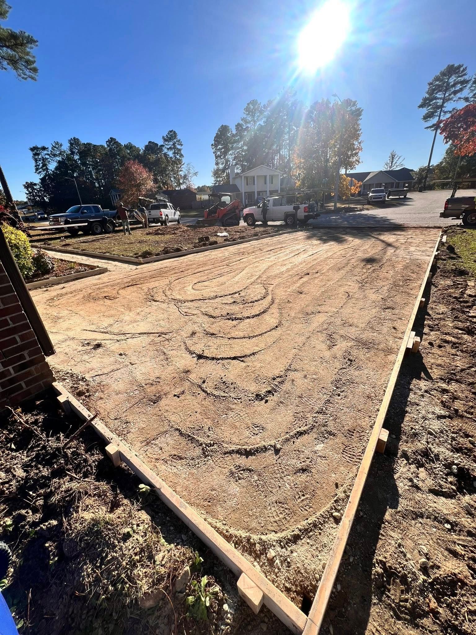 Freshly graded dirt lot under construction with wooden edging and bright sunlight; vehicles and trees in background