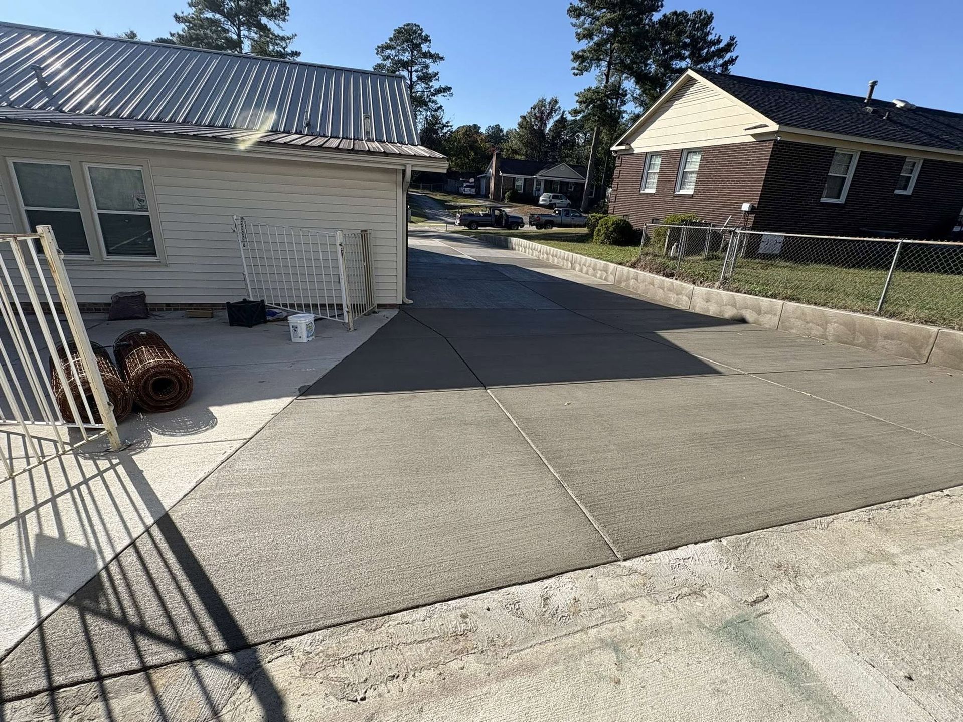 Concrete driveway beside a white house, with a brick building and chain-link fence in the background.